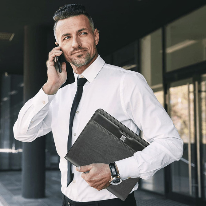 A man in a white shirt and tie holds a black leather zippered compendium while on a phone call.