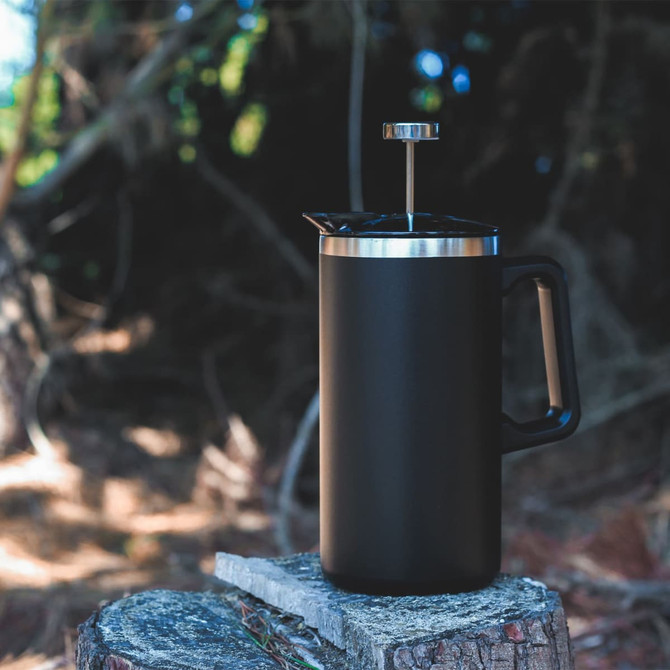 A black coffee press with a stainless steel lid and handle, resting on a wooden stump in a forested setting.