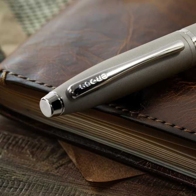 A sleek grey ballpoint pen rests on a leather notebook, featuring a logo on its side.