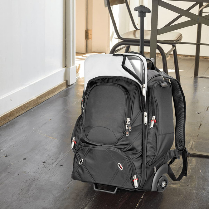 A black wheeled backpack with multiple compartments sits beside a table, featuring a retractable handle and wheels.