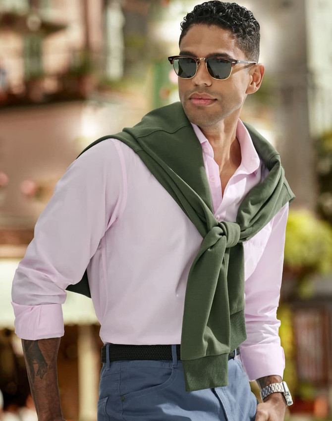 A man wearing a pink shirt and sunglasses, with a green sweater tied around his shoulders, poses outdoors.