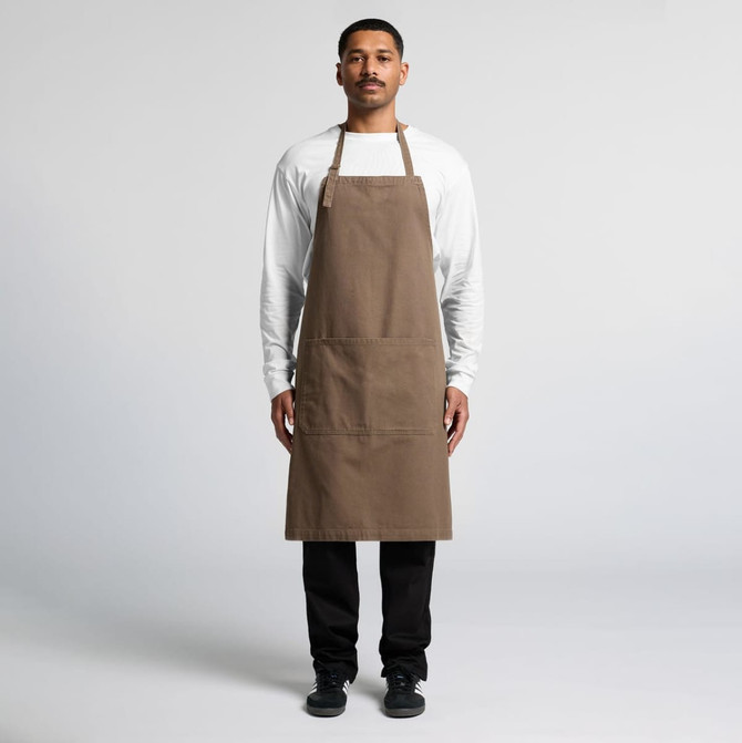 A man stands wearing a brown canvas apron over a white shirt, featuring a logo and front pockets.