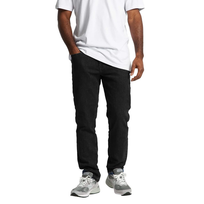 A pair of black jeans displayed on a person wearing a white shirt and grey sneakers. The jeans have a logo.