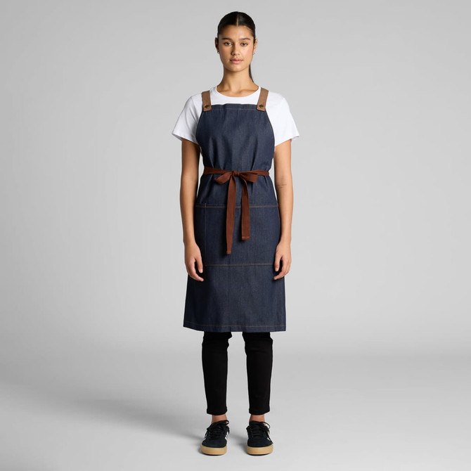 A denim apron featuring brown leather straps and a tie, displayed on a model against a neutral background.