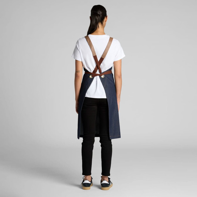 A denim apron with brown straps, worn by a person facing away, paired with a white t-shirt and black trousers.