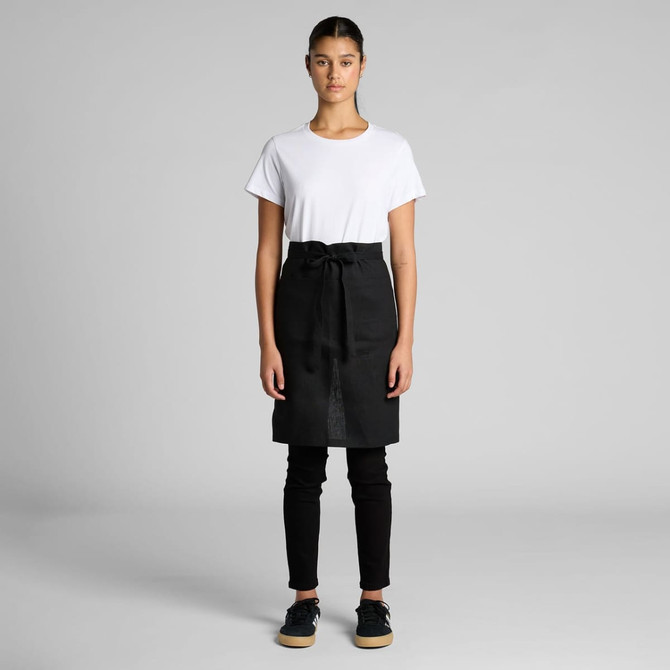 A linen half apron in black is worn over a white t-shirt and black trousers, featuring a tied waist.