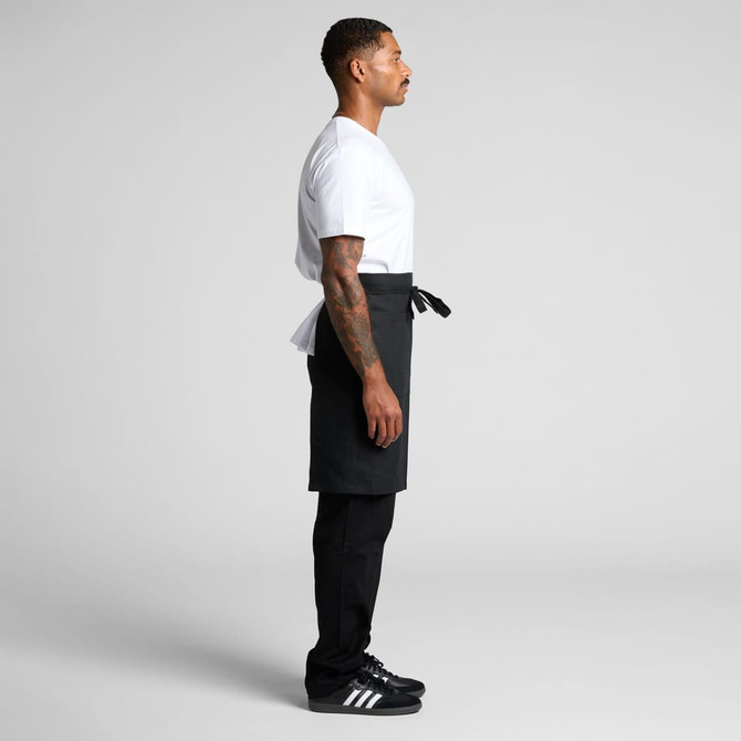 A man stands sideways wearing a black linen half apron over black pants and a white t-shirt.