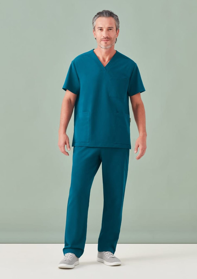 A man wearing unisex teal scrub pants and a matching scrub top, standing against a light green background.
