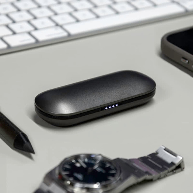 A black charging case for true wireless earbuds rests on a desk next to a watch, a smartphone, and a pen.