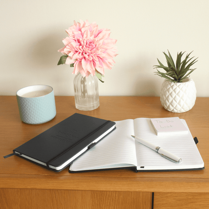 A Pineapple Leather Bound JournalBook in black, opened with a pen, alongside a flower vase and potted plant on a wooden surface.