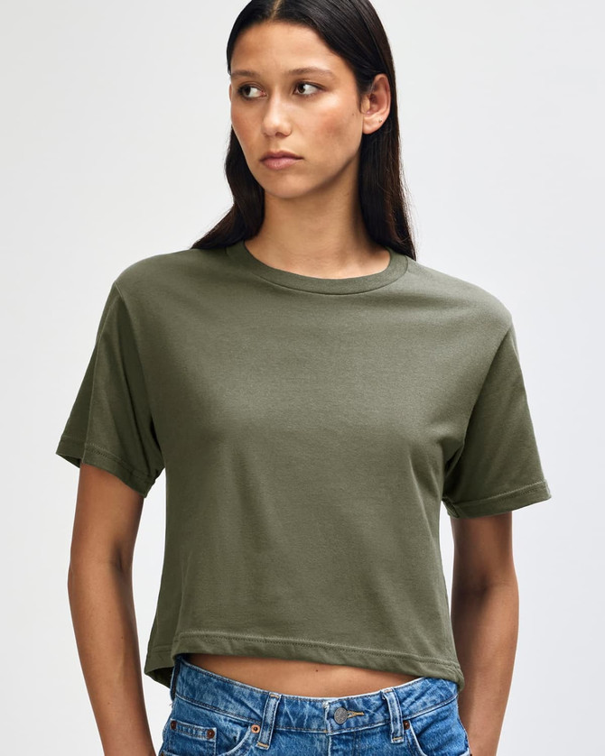 A woman in an olive green boxy T-shirt, styled with high-waisted jeans and curly hair. The shirt has a logo.