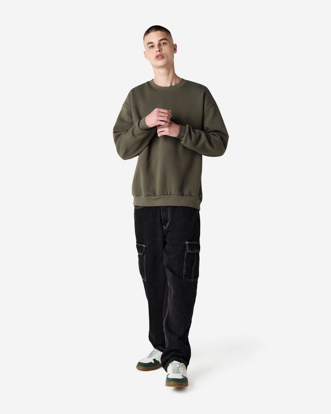 A unisex crewneck sweatshirt in olive green, paired with black cargo pants, worn by a model. Features a logo.