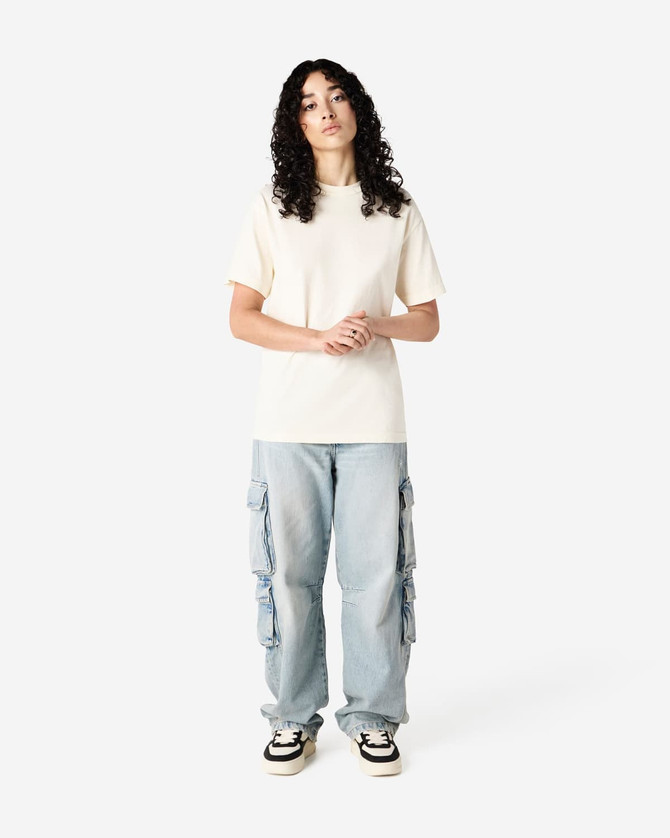 A unisex heavyweight cotton T-shirt in cream, paired with blue cargo pants and sneakers. The model has curly hair and a neutral pose.