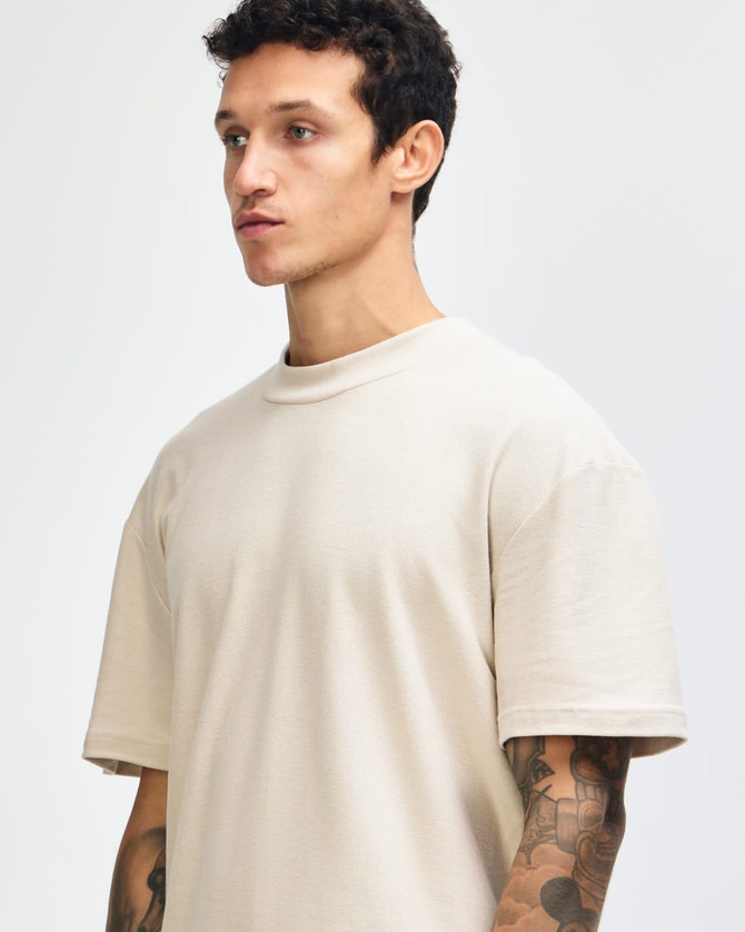 A beige unisex mockneck T-shirt displayed on a model with short sleeves and a relaxed fit. Features a logo.