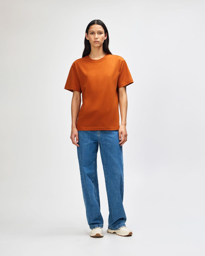 A model wearing an orange heavyweight t-shirt paired with olive green cargo pants and white sneakers, standing against a plain background.