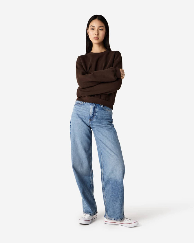 A woman stands confidently in a brown crewneck sweatshirt and light blue jeans, wearing white sneakers.