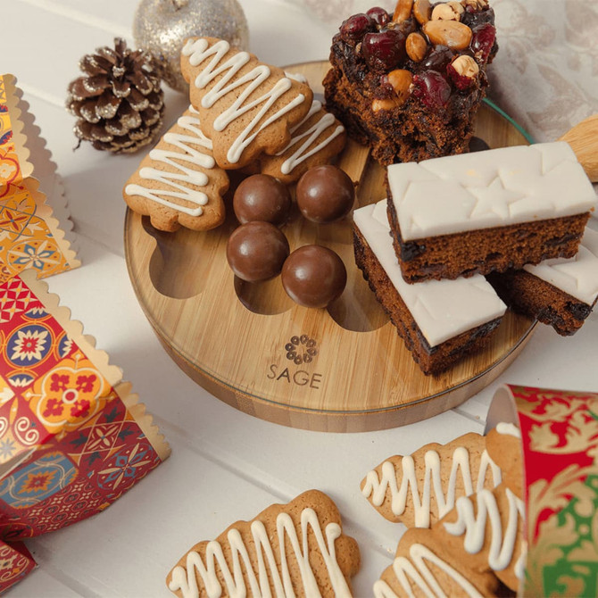 A bamboo platter with decorated gingerbread cookies, chocolate balls, and festive slices, surrounded by wrapping paper and ornaments.