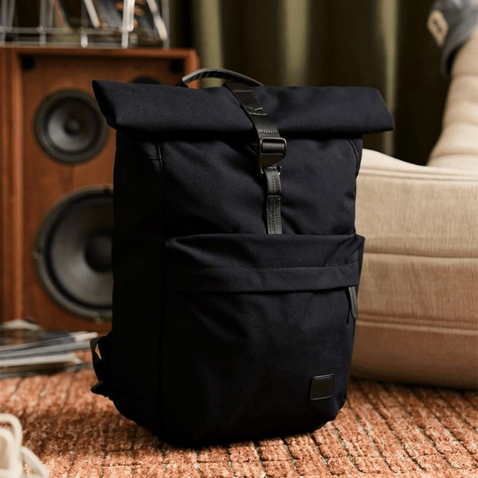 A black rolltop backpack with a front pocket and a branding logo, set against a stylish indoor backdrop.