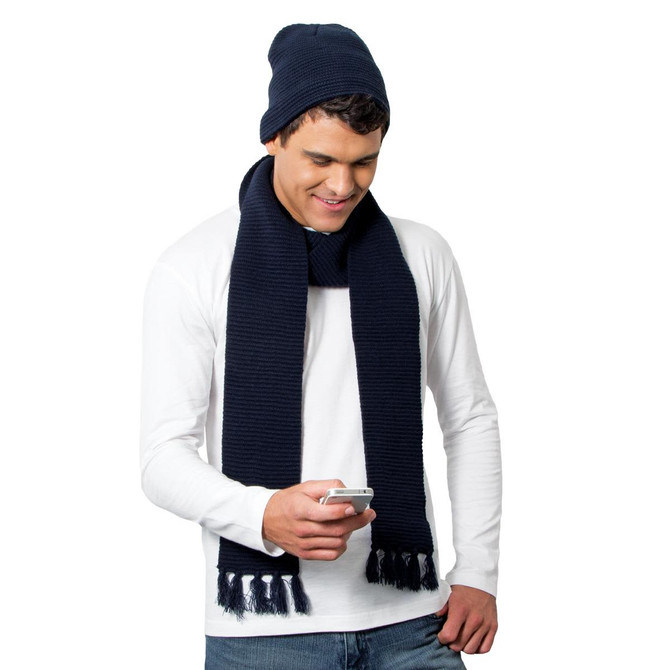 A man wearing a navy Ruga Knit Beanie and matching scarf, holding a smartphone while smiling.