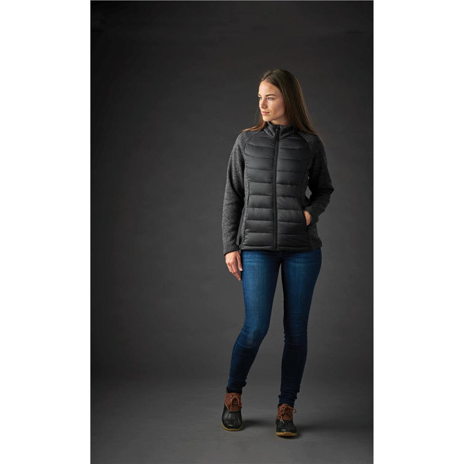 A woman stands wearing a black and grey women's hybrid jacket, paired with blue jeans and brown boots.