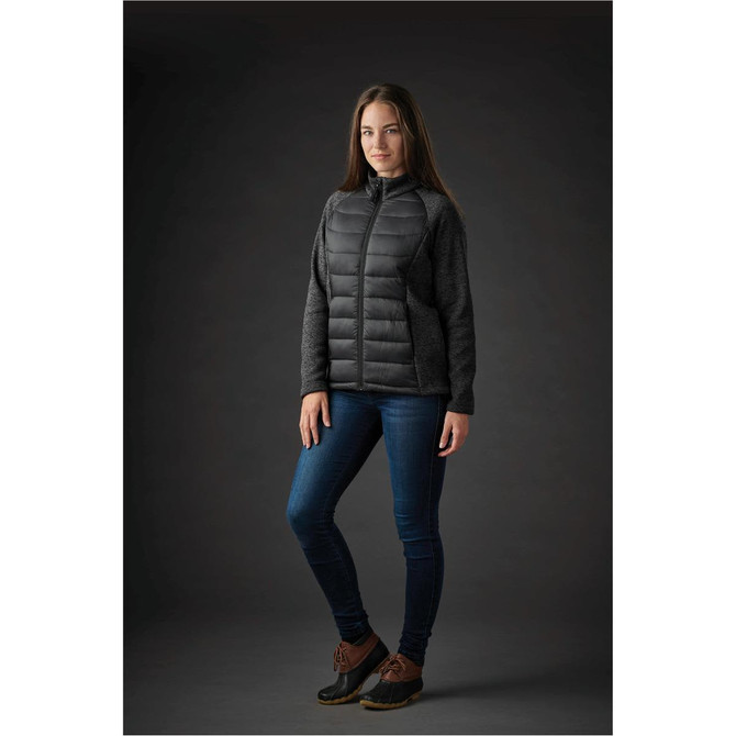 A woman wearing a black and grey Women's Narvik Hybrid Jacket with a fitted design, paired with jeans and boots.