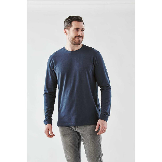 A man wearing a navy long sleeve tee. The shirt features a logo and is paired with grey jeans.