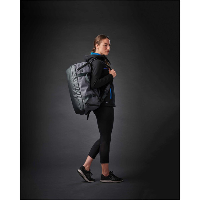 A woman wearing a black jacket and leggings carries a grey duffel bag on her back, set against a dark background.