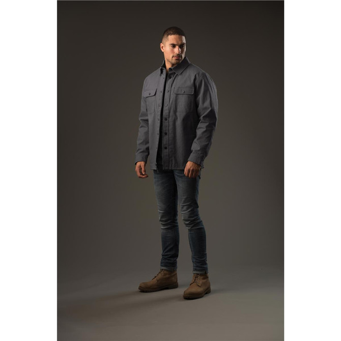 A man stands wearing a grey Men's Tradesmith Jacket, styled with fitted jeans and tan boots. The background is neutral.