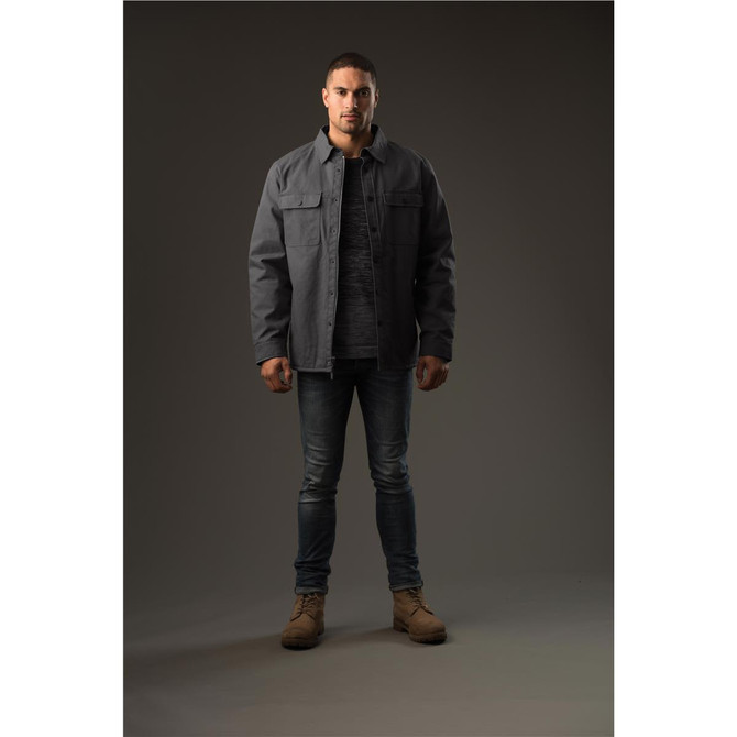 A man stands wearing a grey Men's Tradesmith Jacket over a grey top, paired with jeans and tan boots.