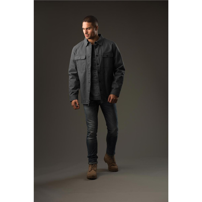 A man wearing a dark grey Tradesmith jacket over a shirt, paired with jeans and tan boots, standing against a neutral background.