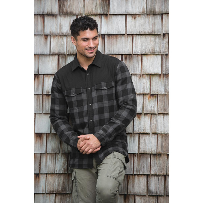 A man wearing a black and grey plaid button-up thermal shirt, standing against a wooden backdrop. The shirt has two front pockets.