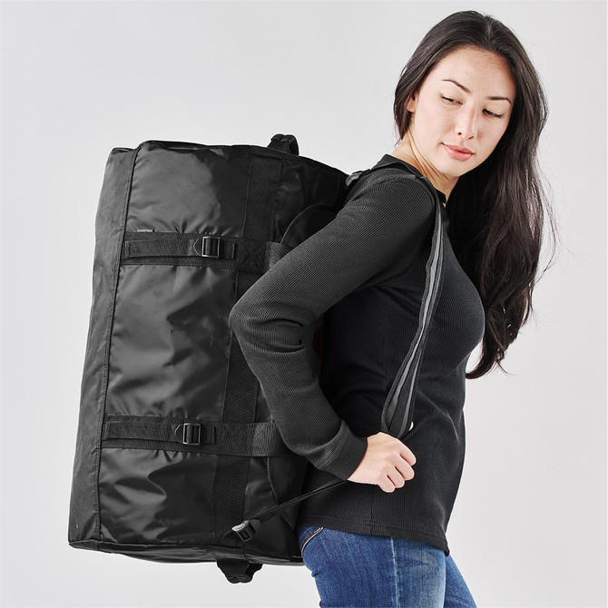 A medium black duffle bag featured being carried by a person, with adjustable straps and buckles visible.
