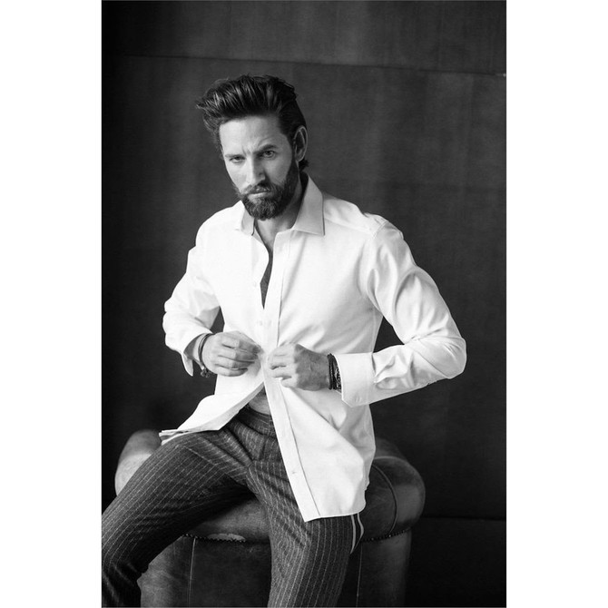 A man in a white button-up shirt sits on a leather stool, adjusting the shirt's buttons. The background is dark.