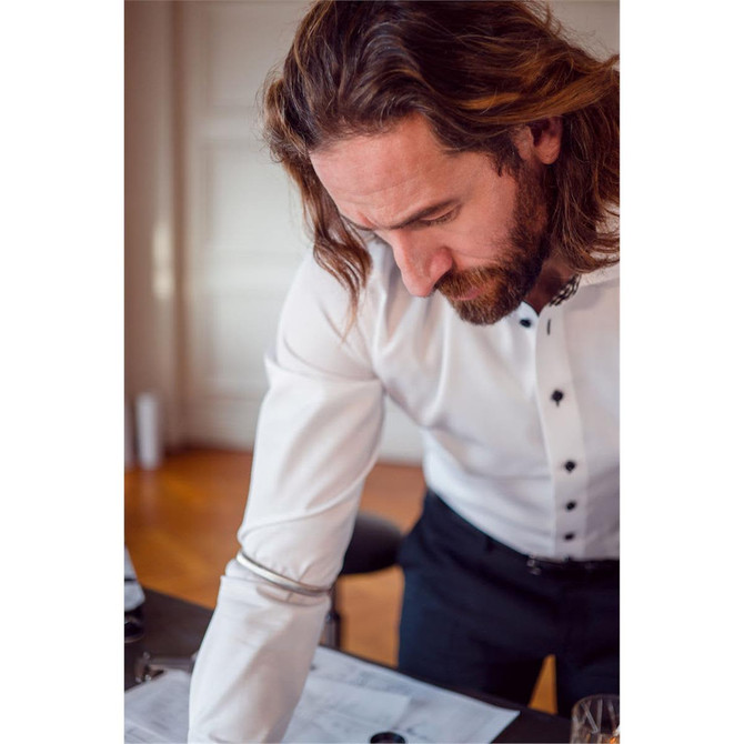 A man in a white button-up shirt with black buttons leans over a table, working on documents.