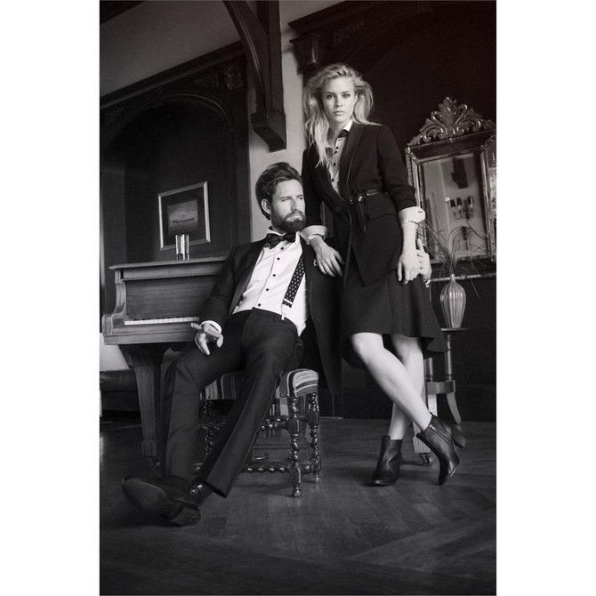 A black and white image of a man in a suit and a woman in a stylish dress, posed elegantly near a piano.