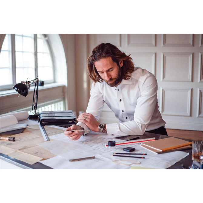 A man with long hair wearing a white button-up shirt, sitting at a desk with architectural plans and tools.