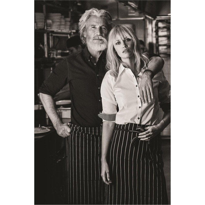 A man with a beard and a woman pose together, both wearing button-up shirts and striped aprons in a kitchen setting.