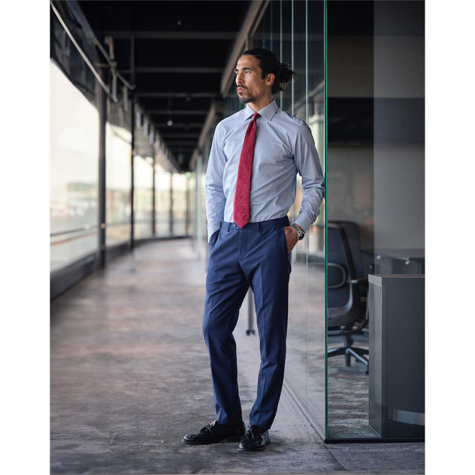 A man in a light blue button-up shirt with a red tie and navy pants stands in a modern office setting.
