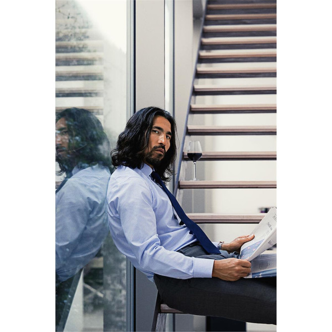 A man with long hair sits on a chair, wearing a light blue button-up shirt and a tie, holding a newspaper.