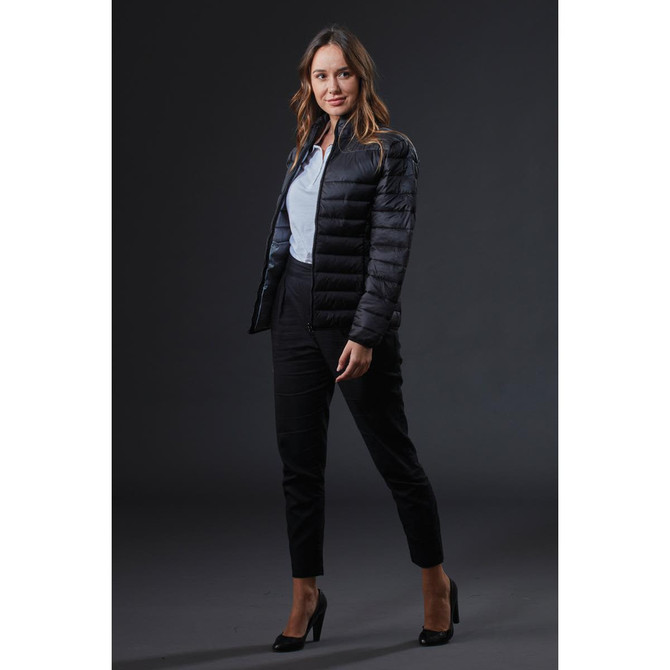 A woman in a black puffer jacket poses against a dark background, wearing tailored black trousers and high heels.