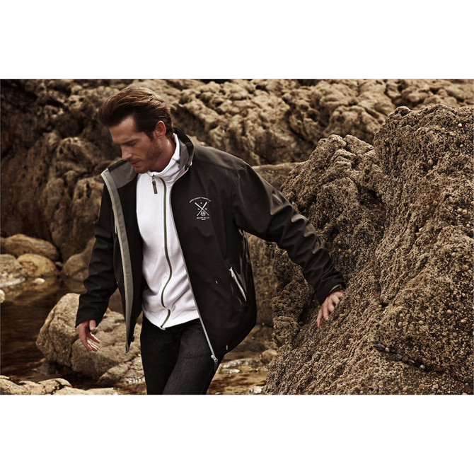 A man wearing a black softshell jacket with a logo, walking along a rocky coastline.