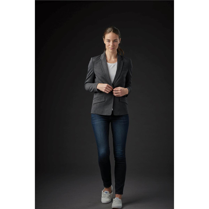 A woman in a grey knit blazer, white top, and jeans, standing against a dark background. The blazer has a logo.