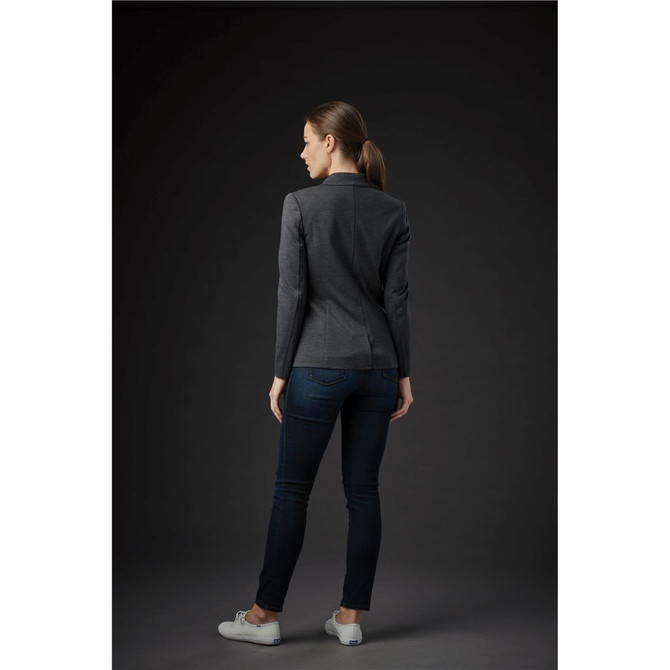 A woman wearing a dark grey knit blazer, paired with dark jeans and white shoes, facing away from the viewer.