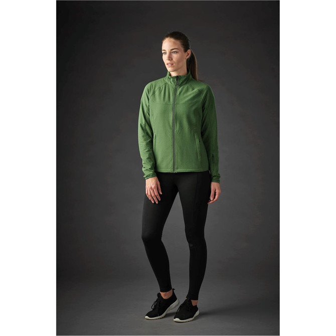 A woman wearing a green jacket with a zip and black leggings, standing against a dark background. The jacket has a logo.