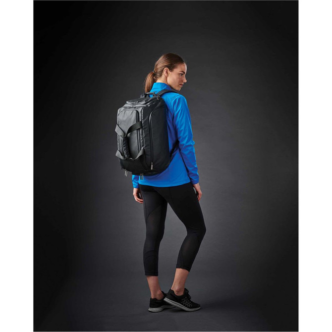 A woman wearing a blue lightweight softshell jacket, black leggings, and a black backpack, standing against a dark background.