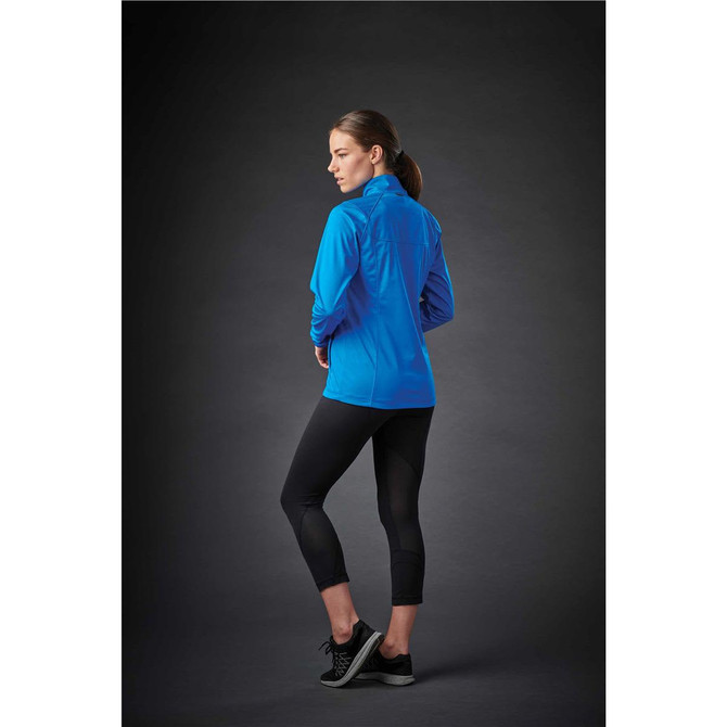 A woman in a blue softshell jacket stands with her back to the camera, wearing black leggings and sneakers.
