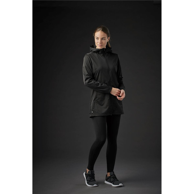 A woman in a black softshell jacket stands against a dark background, paired with black leggings and shoes.