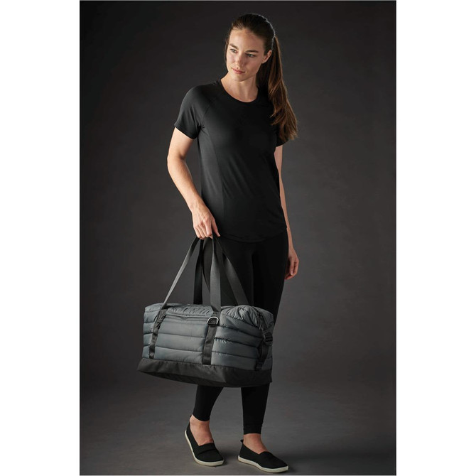 A quilted grey duffle bag is held by a woman wearing a black top and leggings, featuring sturdy handles.