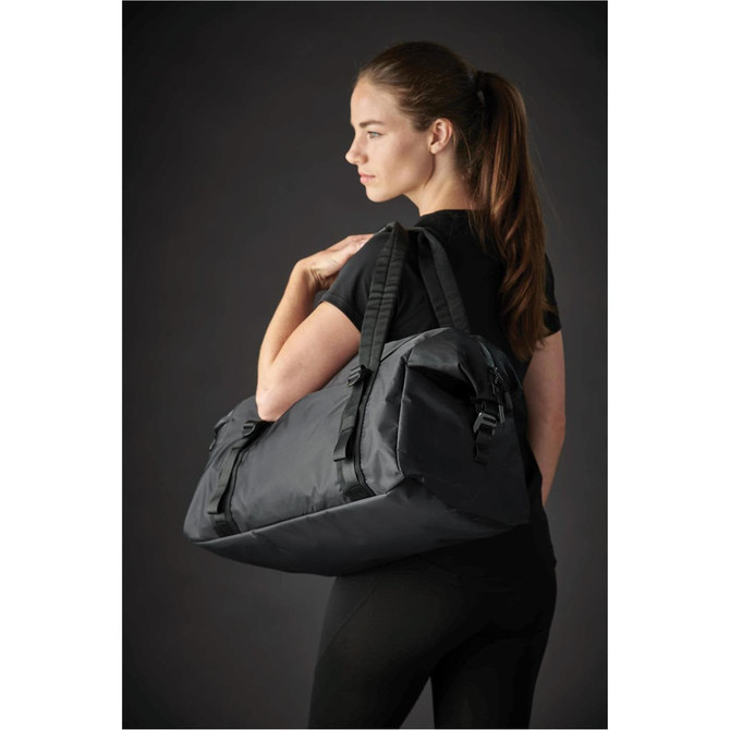 A black duffle bag is worn over the shoulder by a model dressed in black activewear. The bag features a logo.