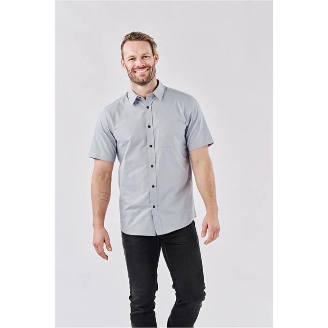 A man wearing a short-sleeved, light blue button-up shirt with a chest pocket and a logo.